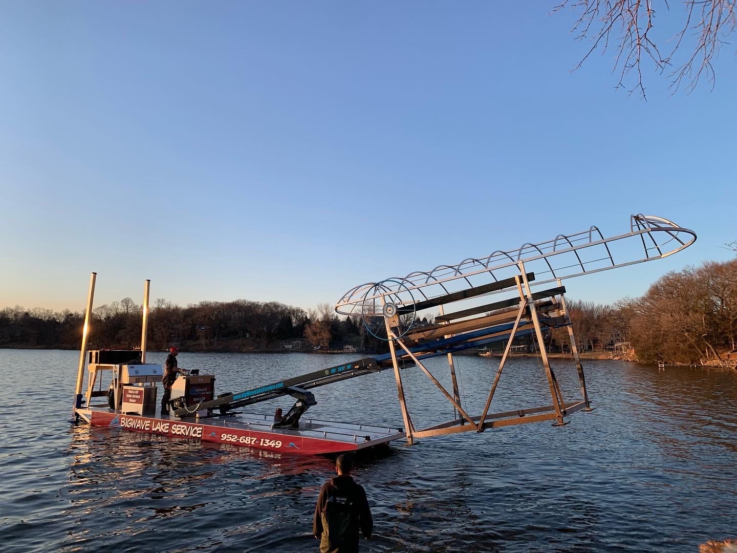 BigWave crew member standing in the lake in waders at dusk while the barge holds a boat lift frame