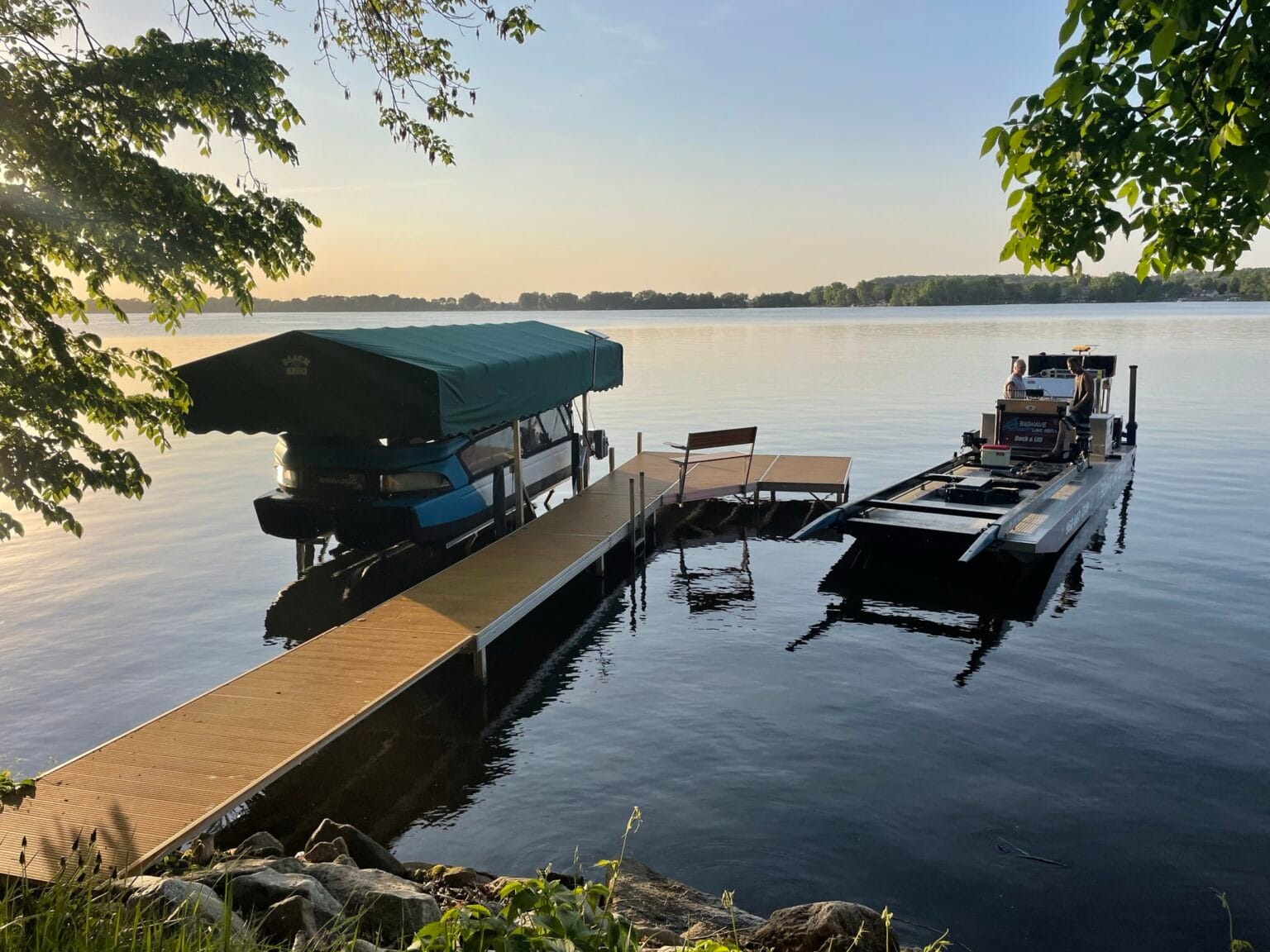 Aluminum dock with tan decking, a green-canopied boat lift and a work barge on a calm lake at dusk