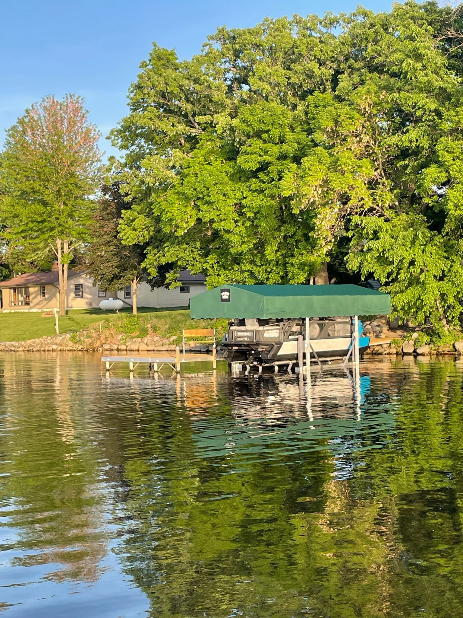Boat lift under a green canopy reflected in still water, in front of a lake home and tall trees
