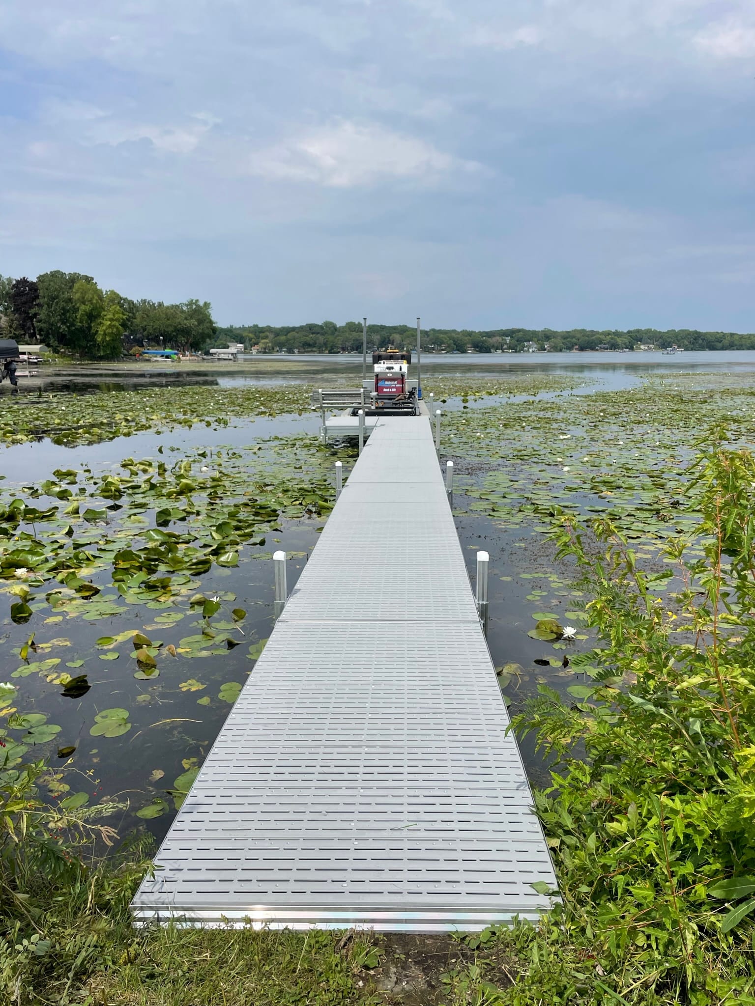 Long aluminum dock running straight out through lily pads to open water, with the BigWave barge at the far end