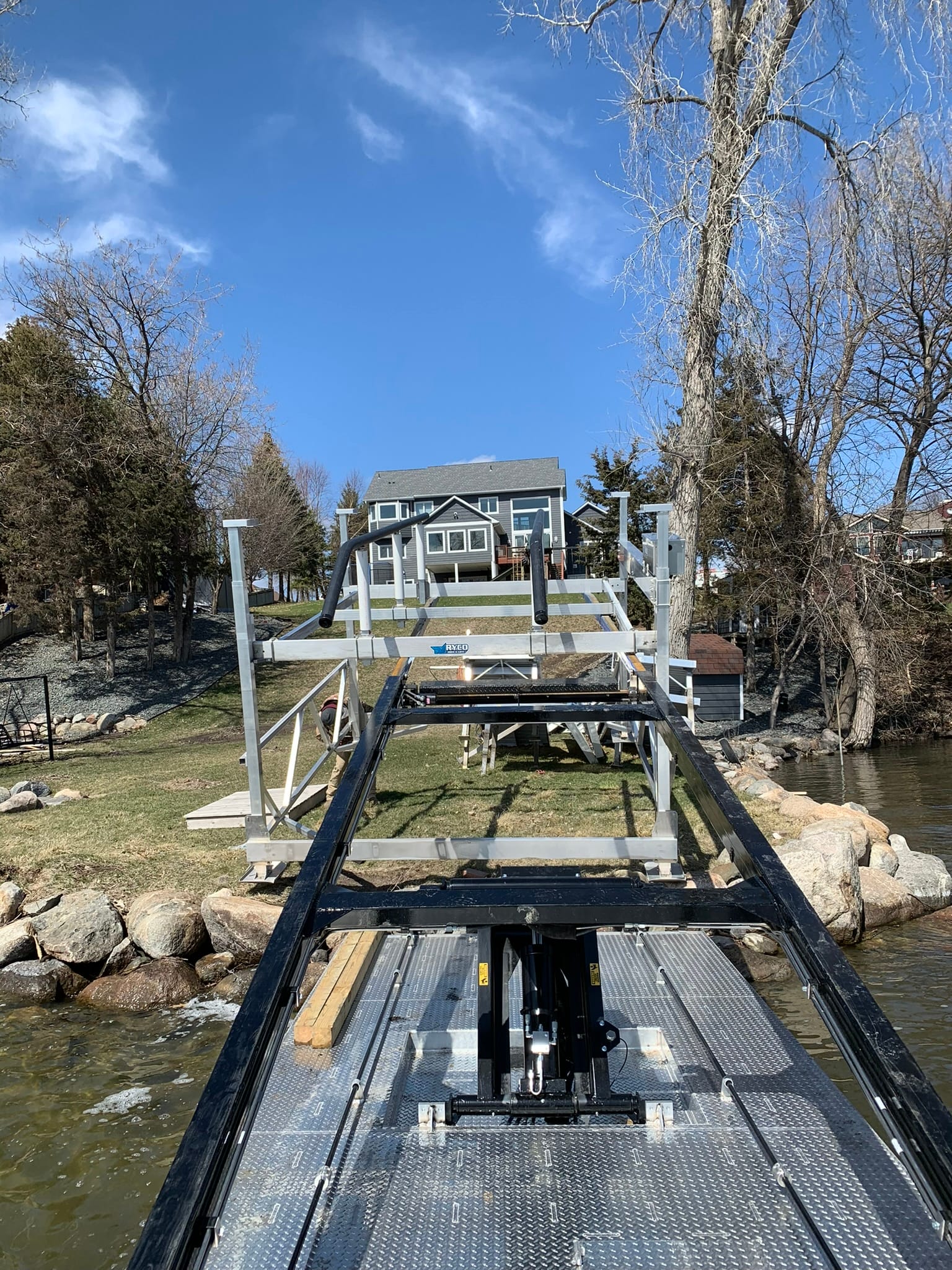 The BigWave barge holding a boat lift frame over a rock shoreline below a lake home, before the trees leafed out