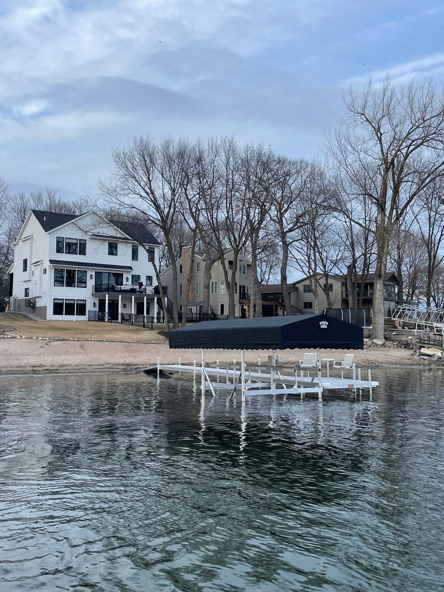 Boat lift and dock sections standing in the shallows in front of a white lake home before the trees leafed out, with a dark canopy cover on the beach