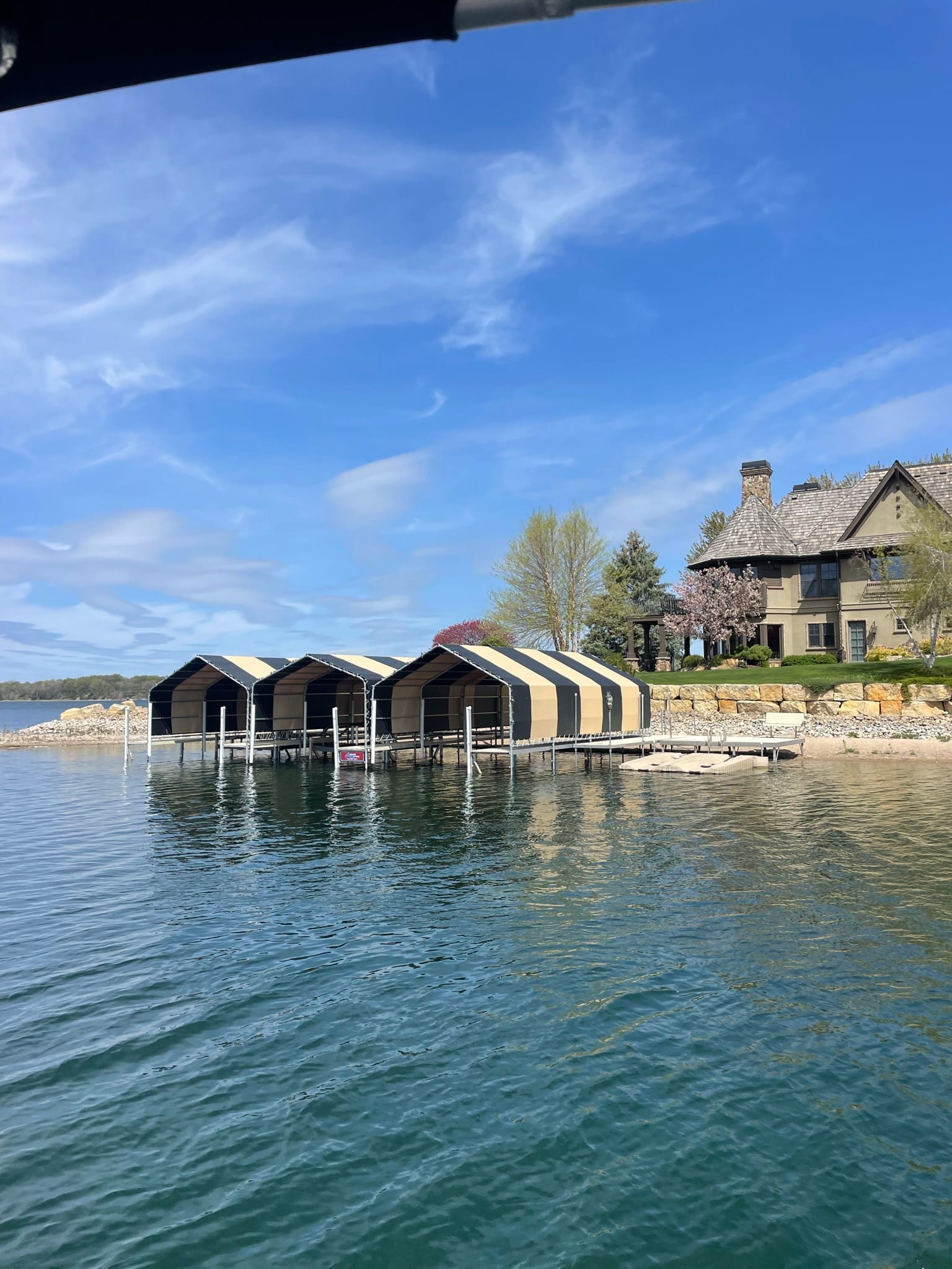 Three boat lifts under black and tan canopy covers in front of a stone lake home