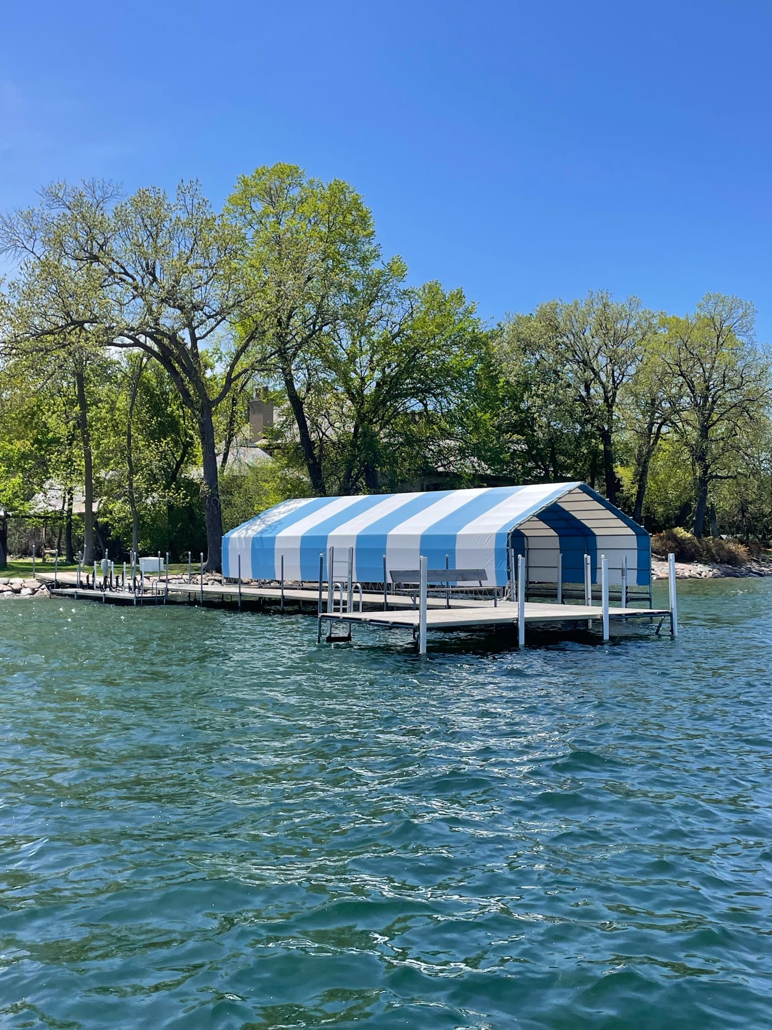Boat lift with a blue and white striped canopy beside a dock, with the shoreline trees in full leaf
