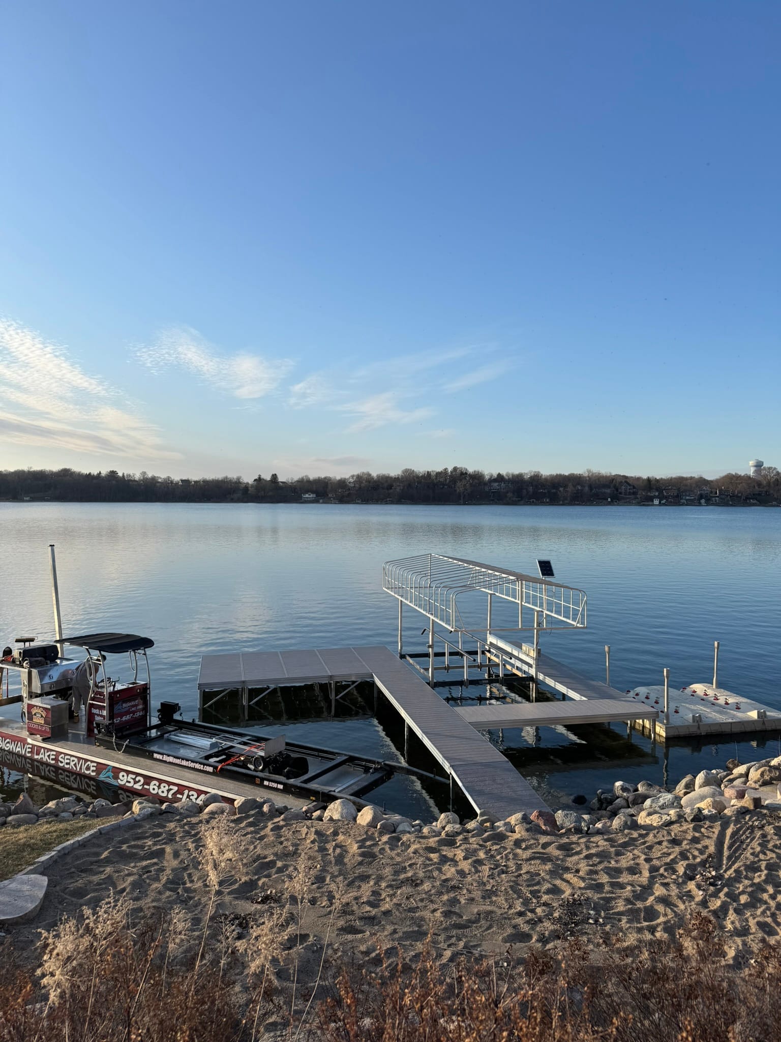 The BigWave barge tied up beside a dock and a lift frame in low sun, with a sandy shore in front