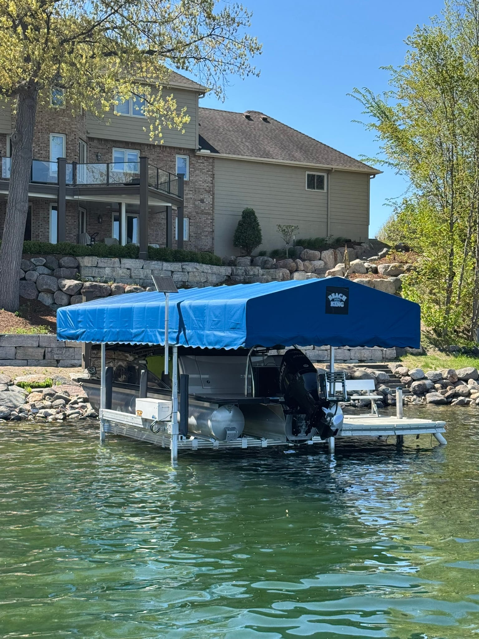 Pontoon on a boat lift under a blue canopy in front of a lake home with a boulder wall