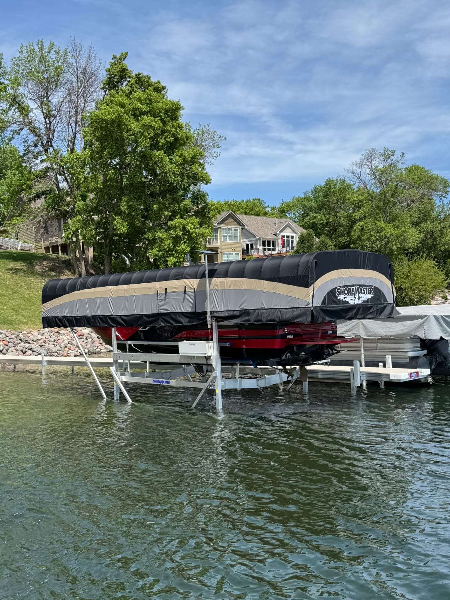 Boat on a lift under a black and tan canopy cover in front of a lake home in summer