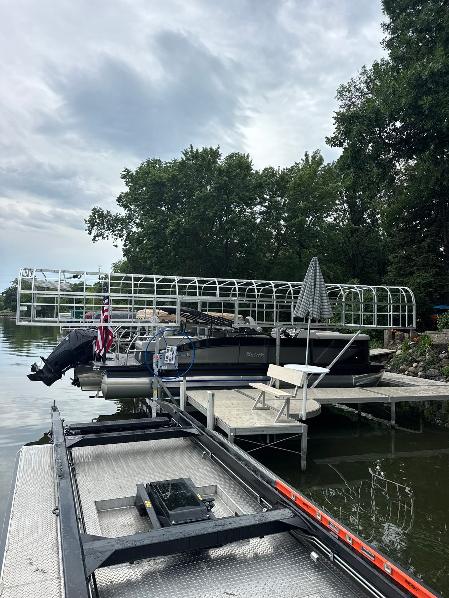 From the deck of the BigWave barge, a pontoon on its lift under a bare canopy frame beside a lake home's dock