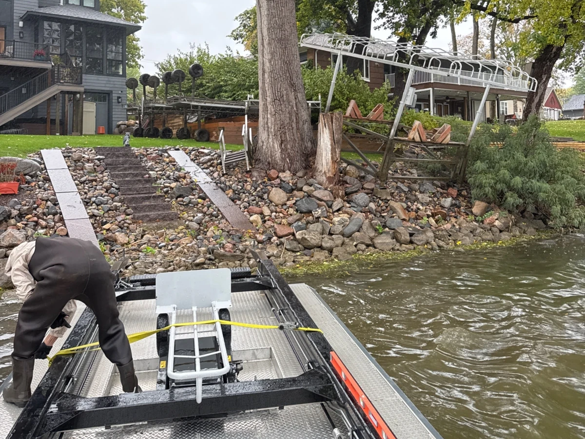 Crew member in waders strapping a boat lift to the deck of the BigWave barge at a rocky shoreline