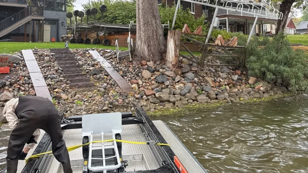 BigWave crew member on the barge at a Spring Lake shoreline after fall removal, with roll-in dock sections and a lift frame pulled up on shore